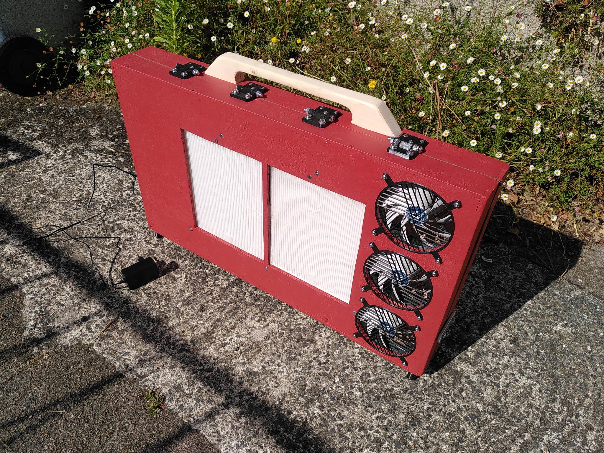 Une valise en bois peinte en rouge, avec une charnière sur le dessus. Sur les grandes faces, deux trous évidés avec des filtres en papier dedans, et trois trous avec des ventilateurs et une grille devant. Une prise électrique sur secteur pour l'alimenter est posée à côté